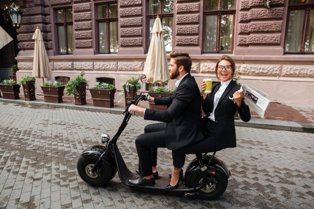 A man and woman in business attire riding a two-person electric scooter on a cobblestone city street, with the woman holding coffee and a newspaper.