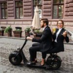 A man and woman in business attire riding a two-person electric scooter on a cobblestone city street, with the woman holding coffee and a newspaper.