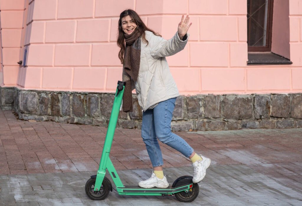Smiling woman in a light jacket and scarf riding a green Lime electric scooter on a paved sidewalk while waving