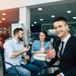 Smiling car salesperson giving a thumbs up as a happy couple receives car keys at a dealership.