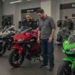 A man and a woman inspecting a red Kawasaki Ninja motorcycle in a showroom. Several other Kawasaki Ninja motorcycles, including a green one in the foreground, are lined up. The showroom features Kawasaki branding, motorcycle gear, and accessories displayed on the walls.