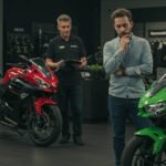 A man in a light blue shirt is thoughtfully examining a green Kawasaki Ninja motorcycle in a showroom. In the background, a sales representative with a notepad observes, and several other Kawasaki Ninja bikes, including red and black models, are displayed. The showroom features Kawasaki branding and motorcycle gear on the walls.