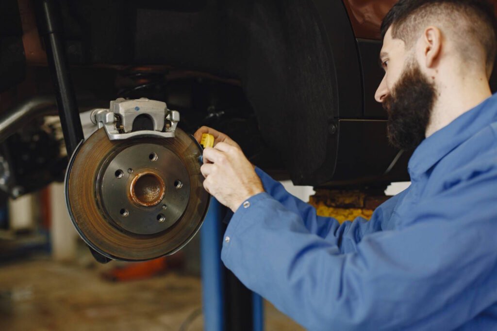 A male mechanic in a blue uniform measuring or inspecting a vehicle’s brake disc and caliper system in an auto repair shop.