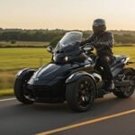 A rider wearing a black leather jacket and helmet is cruising on a sleek black three-wheeled motorcycle along a scenic rural road at sunset. The landscape features lush green fields and a tree-lined horizon, bathed in the warm, golden glow of the setting sun. The motorcycle's aerodynamic design, dual front wheels, and powerful stance highlight its modern engineering and sporty appeal.