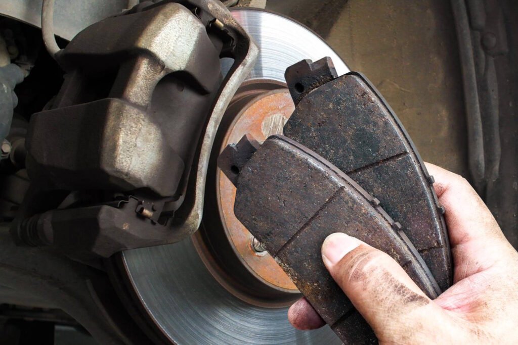 Close-up of a mechanic’s hand holding two heavily worn brake pads near a car’s brake rotor and caliper, indicating the need for replacement.