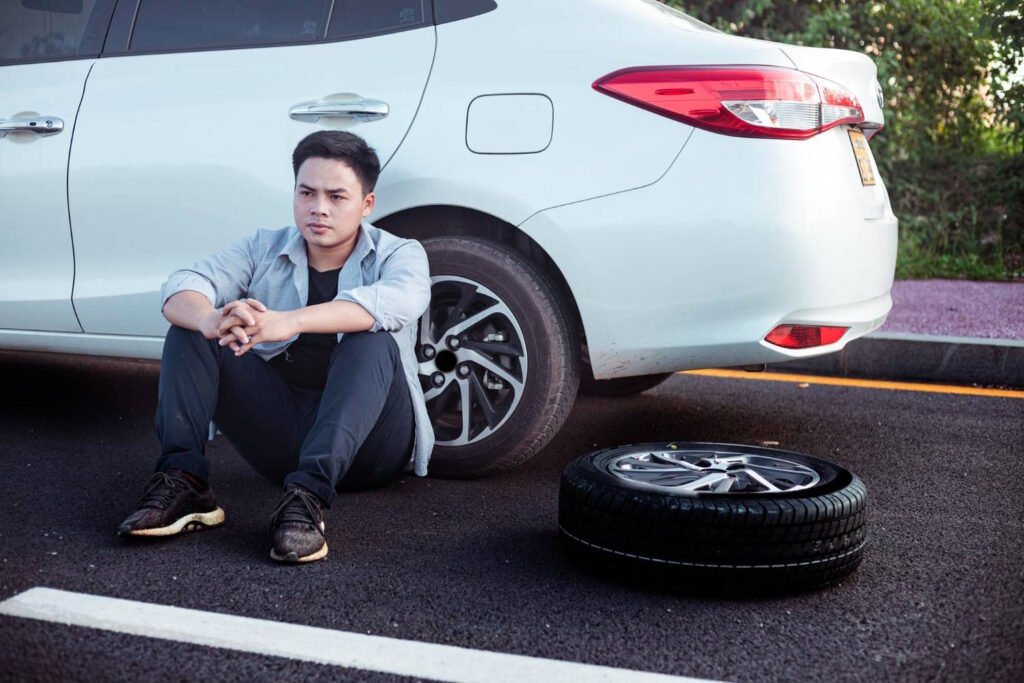 A worried man sits on the roadside next to a white car, concerned about unbalanced tires, with one tire removed and lying on the ground.
