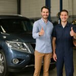 A 2014 Nissan Pathfinder parked in an auto workshop with the smiling car owner and two uniformed mechanics standing beside it, all giving thumbs-up.