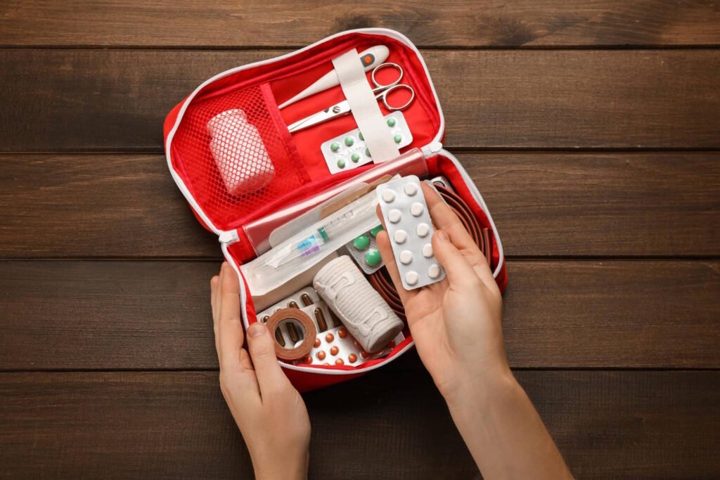 Hands opening a red zippered first aid kit containing pills, bandages, scissors, tape, thermometer, and medical supplies, placed on a wooden table.