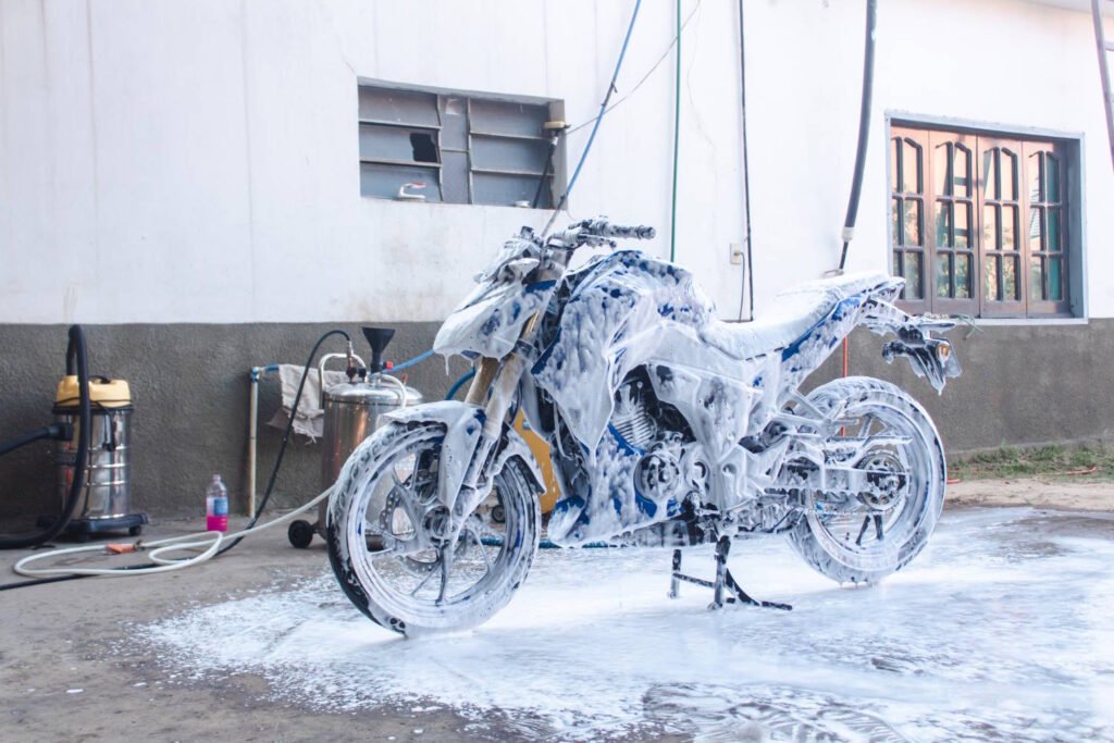 A motorcycle is covered in thick white foam during a wash at home, parked on a concrete surface near a white wall. Cleaning equipment including a vacuum, foam cannon, and hoses are visible in the background, indicating a do-it-yourself motorcycle detailing session.