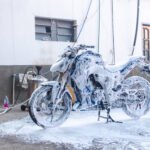 A motorcycle is covered in thick white foam during a wash at home, parked on a concrete surface near a white wall. Cleaning equipment including a vacuum, foam cannon, and hoses are visible in the background, indicating a do-it-yourself motorcycle detailing session.