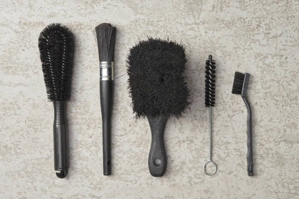 A set of five specialized motorcycle cleaning brushes arranged on a concrete surface. The set includes a wheel brush, detailing brush, sponge-style body brush, narrow crevice brush, and a stiff-bristle brush, ideal for cleaning tight spaces, spokes, engines, and chains.