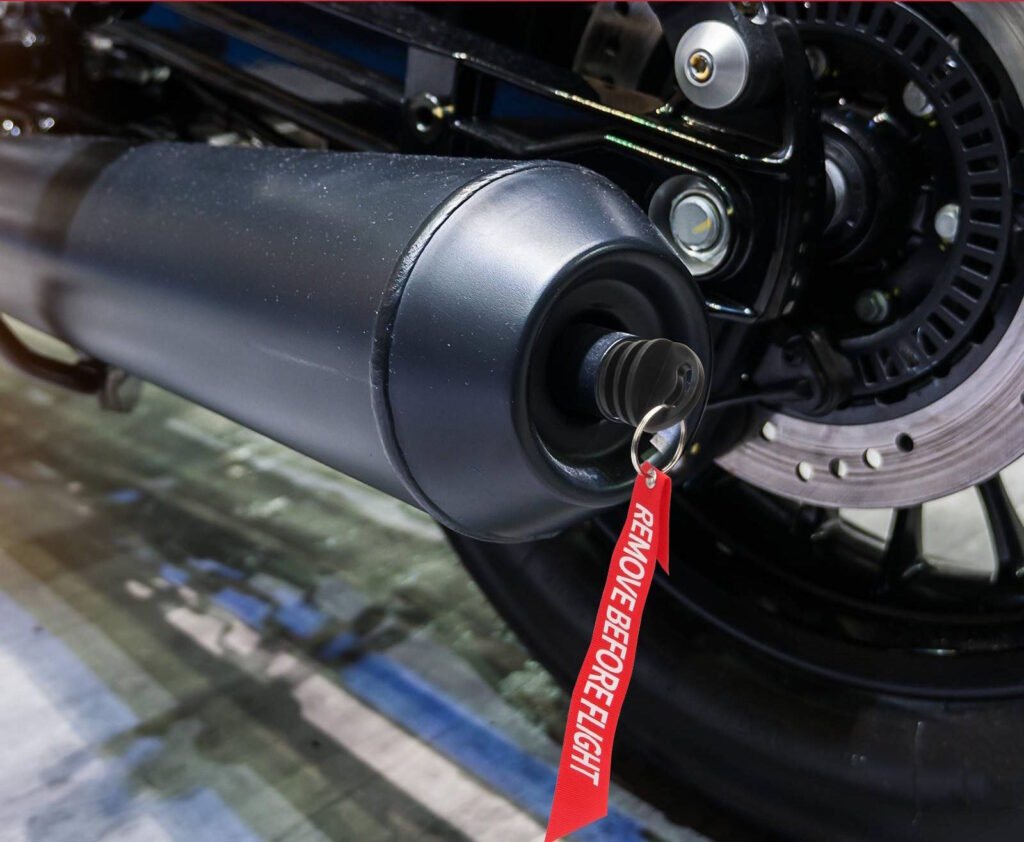 Close-up of a motorcycle exhaust pipe with a rubber exhaust plug inserted, featuring a red “Remove Before Flight” tag. The plug is used to prevent water from entering the exhaust during washing or storage.
