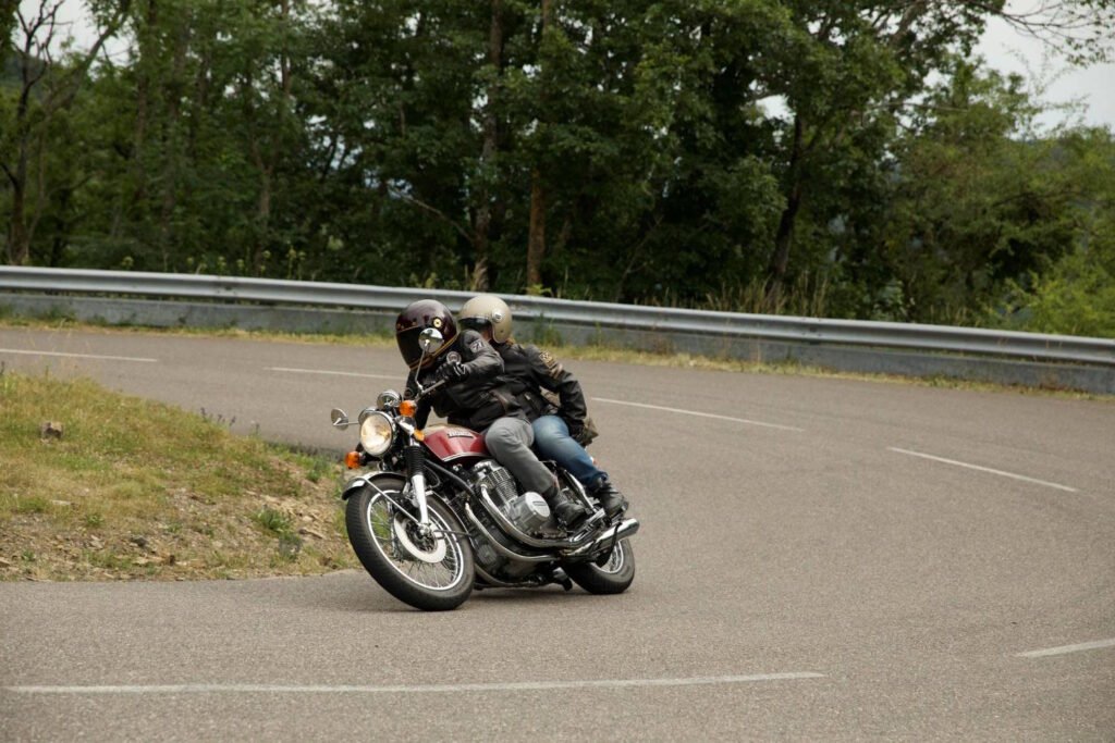 Two motorcyclists in full riding gear leaning into a right-hand curve on a forested mountain road, demonstrating proper cornering posture.
