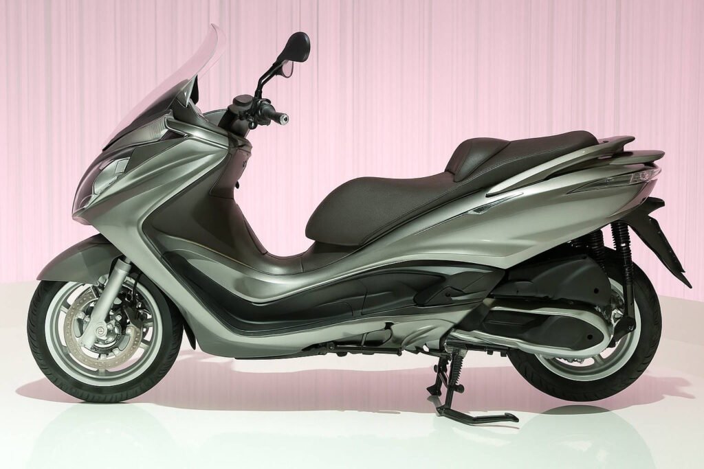 Side view of the Piaggio X10 500 maxi-scooter in silver, displayed on a studio floor with a pink curtain background.