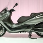 Side view of the Piaggio X10 500 maxi-scooter in silver, displayed on a studio floor with a pink curtain background.