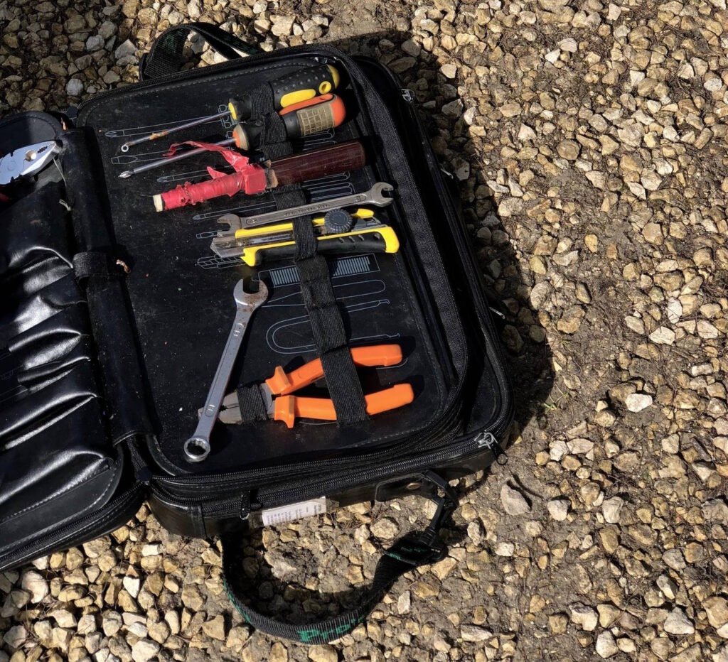 A zippered tool case open on gravel, showing assorted hand tools including screwdrivers, wrenches, pliers, and wire cutters, organized with elastic straps for easy access during roadside motorcycle repairs. 