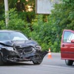 Car accident scene showing a black BMW sedan with significant front-end damage and a red vehicle with its driver's door open, separated by a traffic cone on a road surrounded by trees.