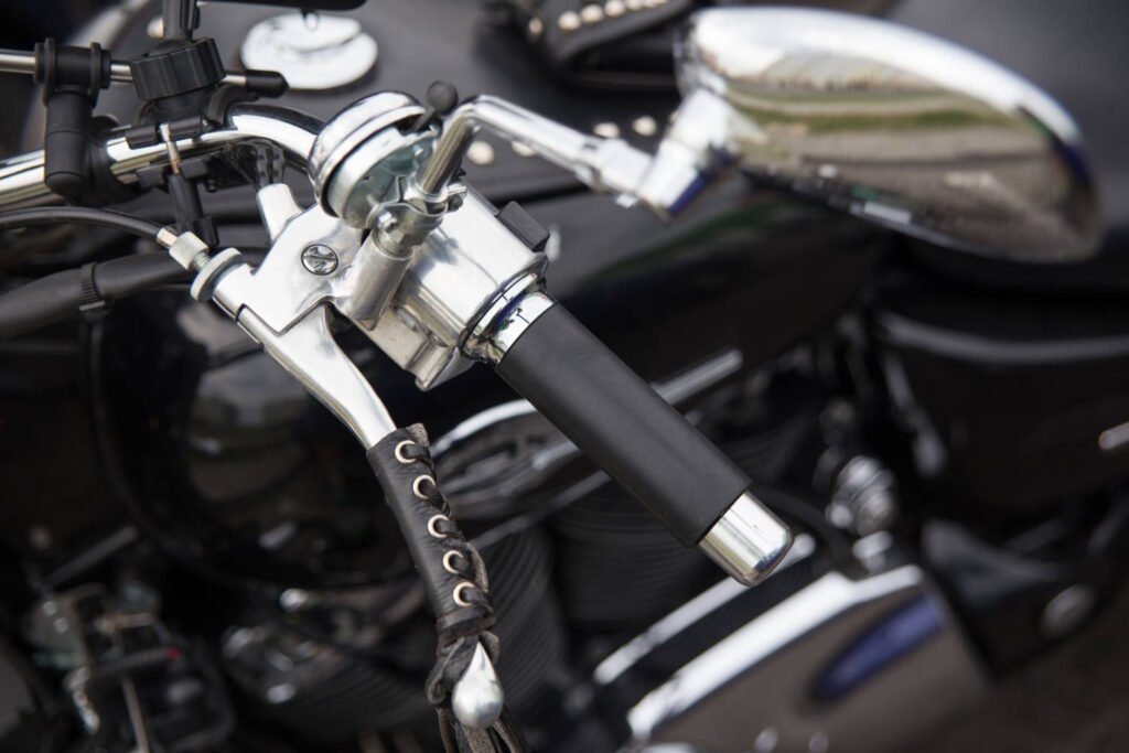 Close-up of a chrome motorcycle clutch lever assembly mounted on handlebars, showing the clutch perch, lever, and barrel adjuster. The handlebar has a black rubber grip and leather wrap detail. Chrome mirrors are visible in the blurred background.