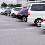 A row of neatly parked cars in a commercial parking lot with clearly marked yellow parking spaces. The vehicles include various models in white, black, and silver colors, all parked in their designated spots with a multi-level parking garage and trees visible in the background.