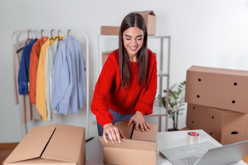 Young woman in red sweater smiling while packing cardboard shipping boxes at desk with laptop, clothing rack with colorful t-shirts visible in background of home office workspace